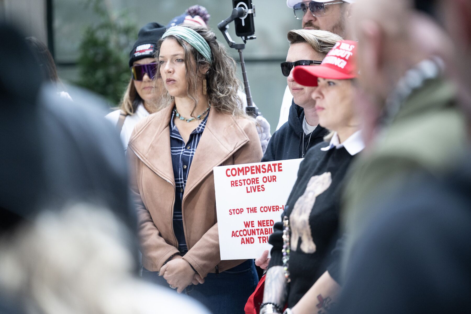People standing together in Washington, D.C., one holding a sign reading Compensate, Restore Our Lives, Stop the Cover-Up